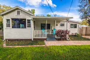 Front facing white bungalow home with small covered porch, pavered walkway and two blue adirondack chairs